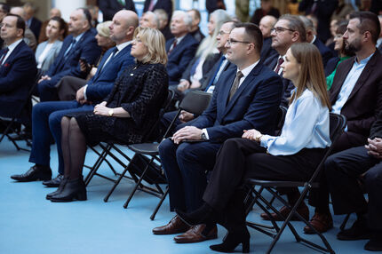 Uczestnicy wydarzenia siedzą w rzędach czarnych składanych krzeseł, ubrani w stroje biznesowe. Wpatrzeni w scenę, słuchają wystąpienia 