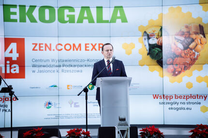 Marszałek w garniturze przemawia z mównicy podczas wydarzenia EKOGALA. W tle widoczny ekran z grafiką promującą Międzynarodowe Targi Produktów i Żywności Wysokiej Jakości i logotypami.