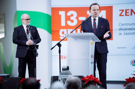 Dwóch mężczyzn w garniturach przemawia na scenie podczas wydarzenia w CWK w Jasionce. Jeden z nich mówi do mikrofonu przy przezroczystej mównicy, drugi stoi obok z rękami splecionymi. W tle ekran z datą 13–14 grudnia i nazwą miejsca.