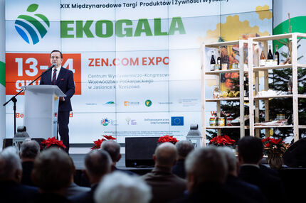 Marszałek przemawia z mównicy podczas wydarzenia EKOGALA, na tle dużego banera promującego Międzynarodowe Targi Produktów i Żywności Wysokiej Jakości. Obok niego stoi drugi mężczyzna. Scenę zdobią choinki i poinsecje, a na banerze widnieje m.in. data
