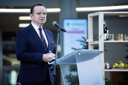 Marszałek w granatowym garniturze, białej koszuli i krawacie w czerwono-niebieskie pasy przemawia przy mównicy z mikrofonem. Ma czerwoną poszetkę w kieszeni. Za nim widoczna jest drewniana półka z produktami spożywczymi — butelkami, słoikami i owocami.