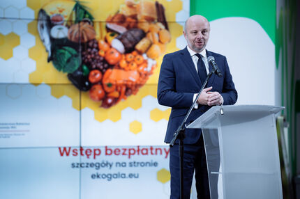 Prezydent w ciemnym garniturze przemawia przy przezroczystej mównicy z mikrofonem, na tle kolorowej grafiki przedstawiającej warzywa, owoce, pieczywo i ryby ułożone w serce na żółto-białym tle przypominającym plaster miodu. W tle widoczne napisy.