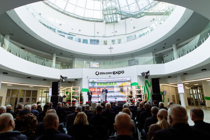 Widok na scenę podczas XIX Międzynarodowych Targów Produktów i Żywności Wysokiej Jakości EKOGALA, odbywających się 13–14 grudnia w nowoczesnym, przeszklonym atrium z kopułą. Dwóch prelegentów przemawia do zgromadzonej publiczności. Tło sceny zdobią banery