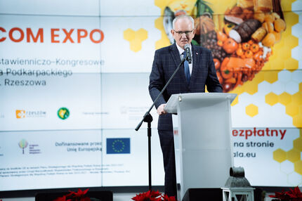 Poseł w garniturze przemawia przy mównicy podczas wydarzenia ECOM EXPO. W tle widoczny jest ekran z nazwą wydarzenia, logotypami instytucji wspierających (w tym Unii Europejskiej) oraz grafiką przedstawiającą żywność — warzywa, owoce, pieczywo 