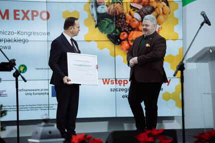 Marszałek oraz Dyrektor ZSG stoją na scenie podczas wydarzenia w Jasionce. Jeden z nich trzyma dużą, oprawioną tablicę z napisem „Kwota pomocy: 40 000,00 zł”. W tle widoczny jest ekran z grafiką przedstawiającą owoce i warzywa w układzie plastra miodu. 