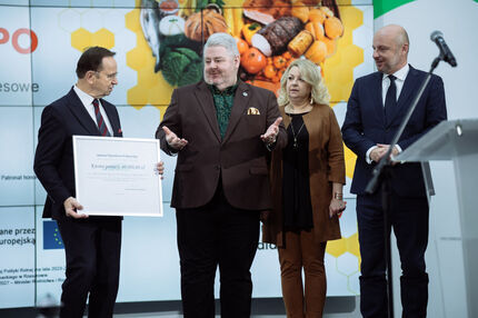 Cztery osoby stoją na scenie podczas wydarzenia w Jasionce. Marszałek trzyma dużą tablicę z napisem „Kwota pomocy: 40 000,00 zł”, informującą o przyznanym wsparciu finansowym. W tle widoczny jest ekran z grafiką przedstawiającą produkty ekologiczne